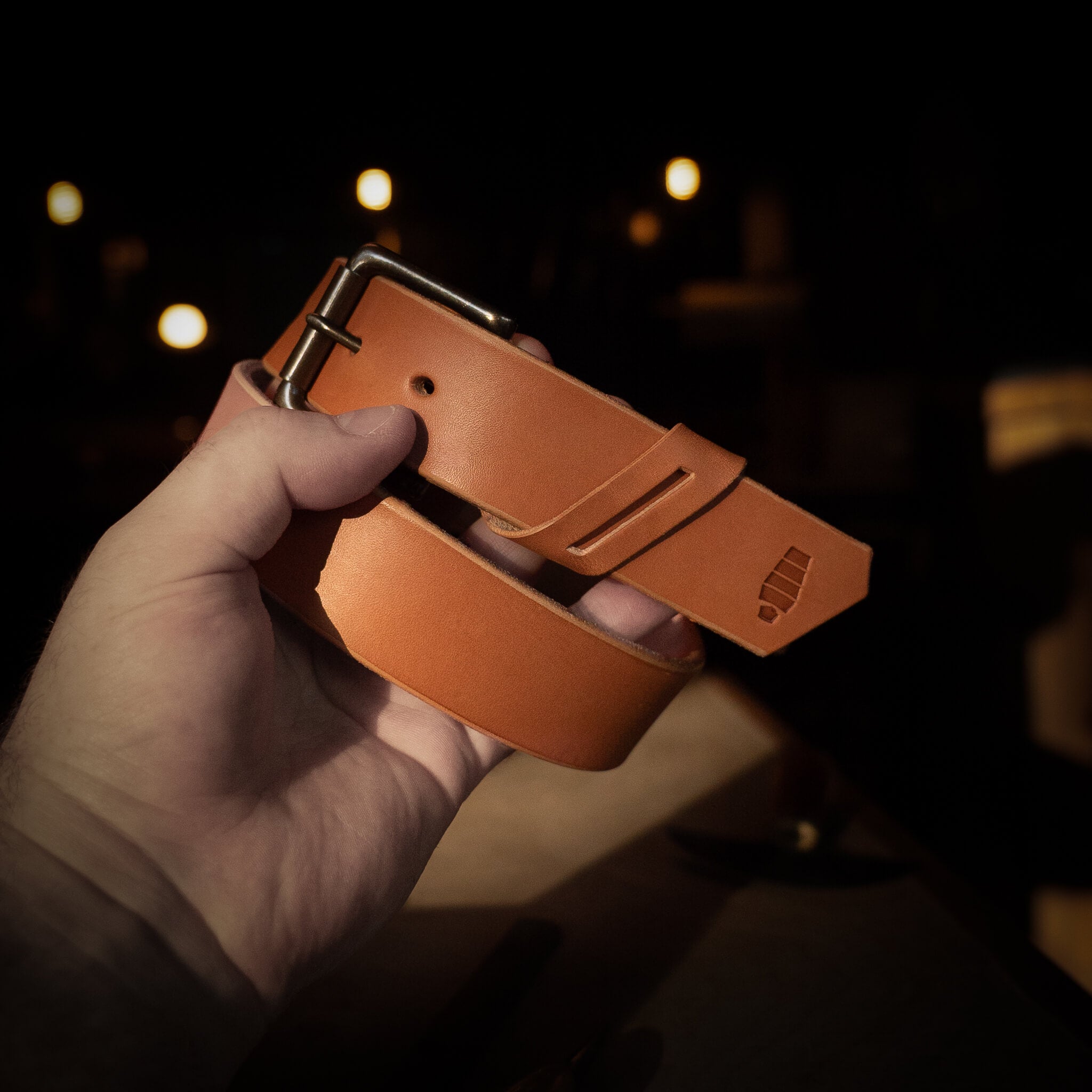 Hand holding a Tan leather belt against a dark background