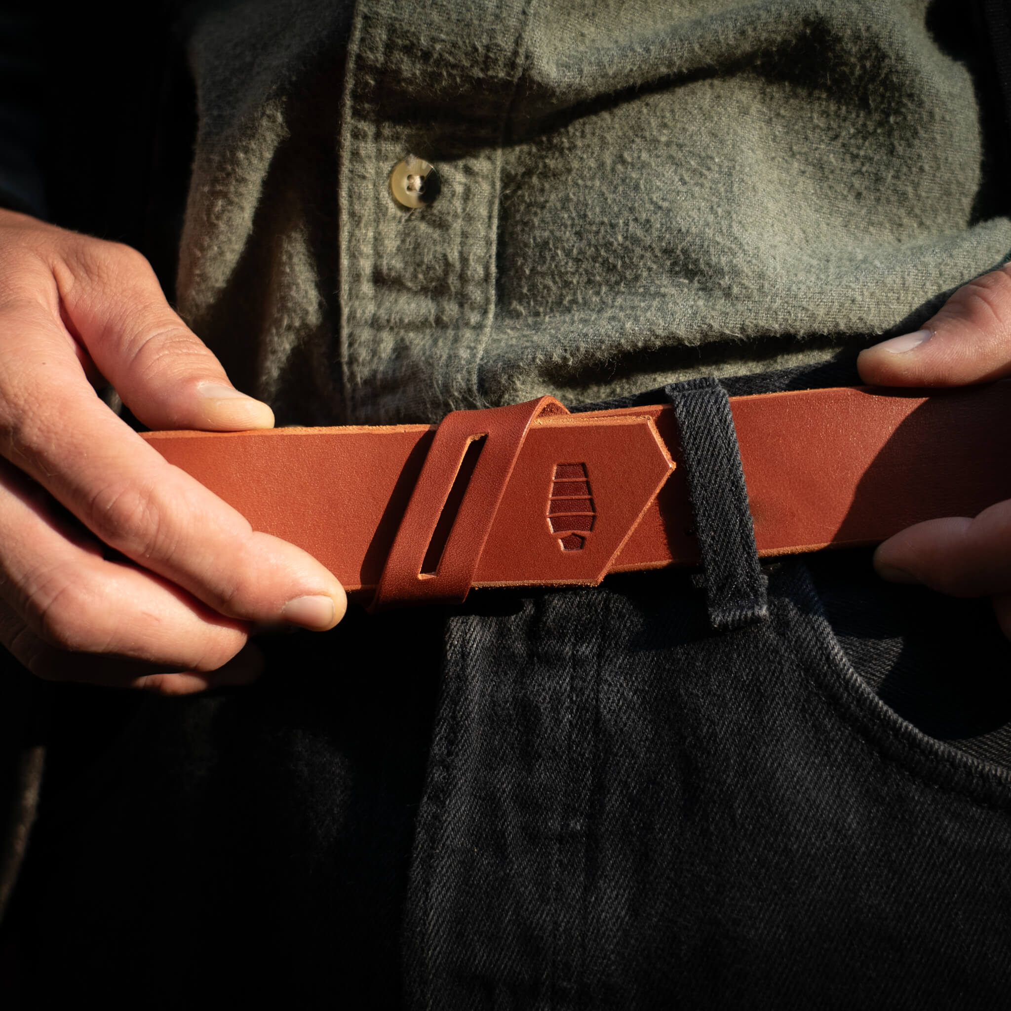 Person threading the tip of a chestnut leather belt into a unique diagonal belt keeper design.
